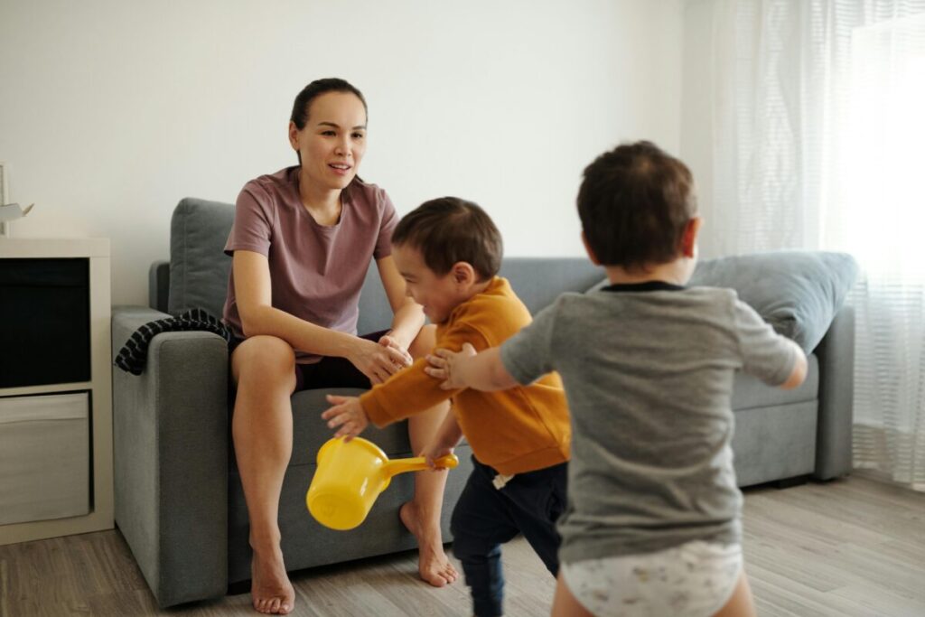 A relaxed mother sitting on a couch with a coffee mug while her young child engages in independent play nearby without mom guilt