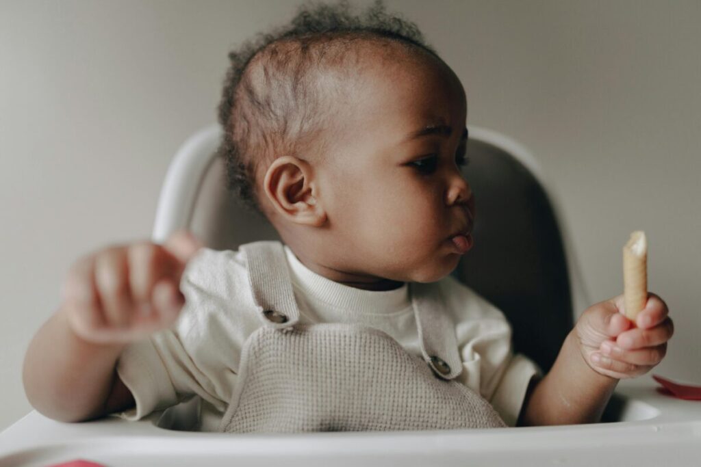 A messy 15-month-old toddler sitting in a high chair and happily self-feeding healthy finger foods, representing a realistic 15 month old feeding schedule for TinyCareTips.