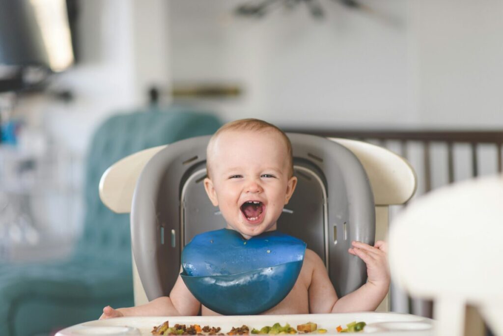 A messy 14 month old toddler eating healthy finger foods in a high chair, practicing his daily feeding schedule.