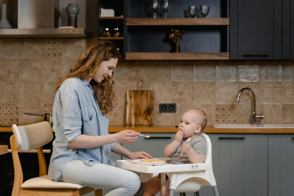 A mother smiling while letting her toddler self-feed safely, practicing toddler food strike at 15 months tips without pressure.